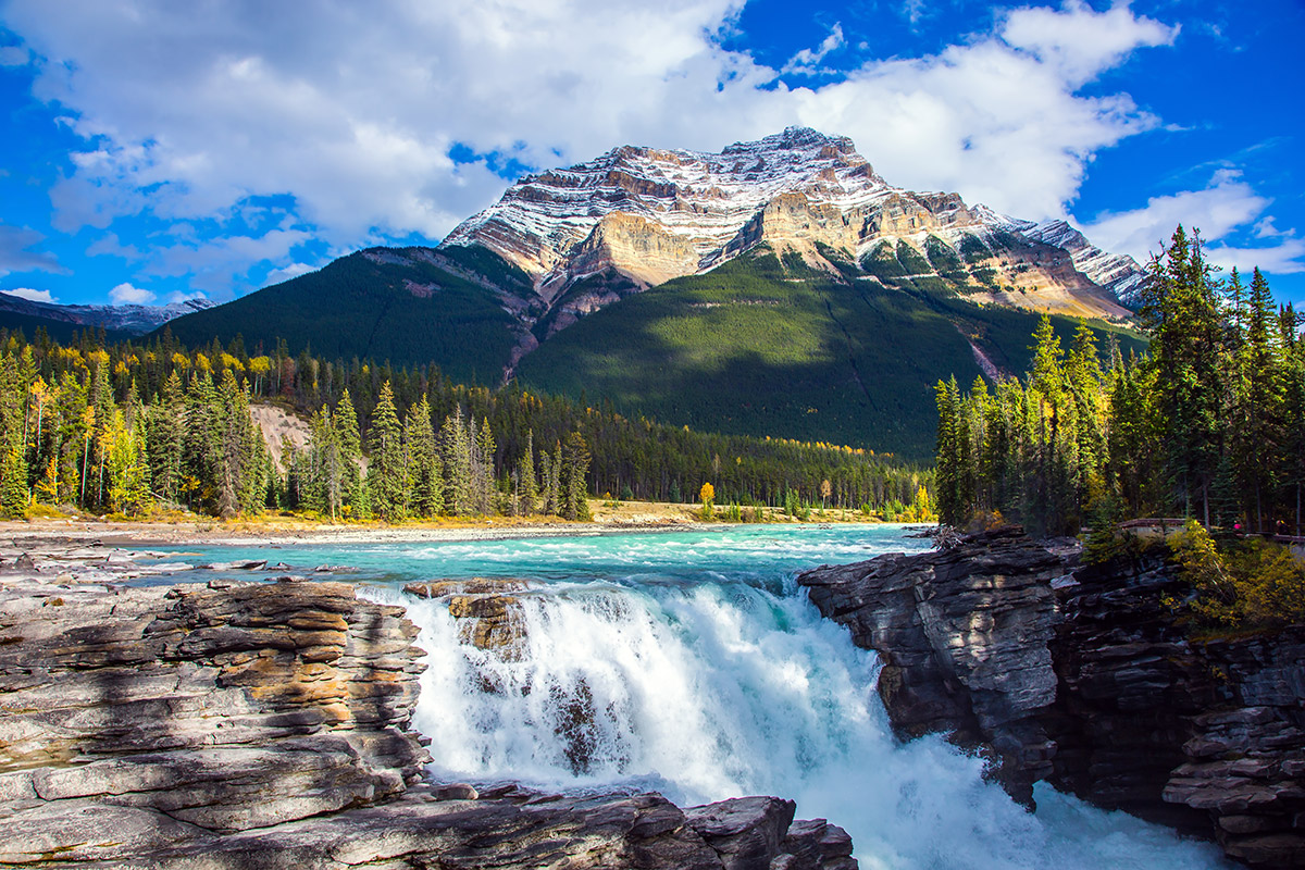 A picturesque waterfall framed by greenery and a backdrop of rocky mountains, under a blue sky with fluffy white clouds.